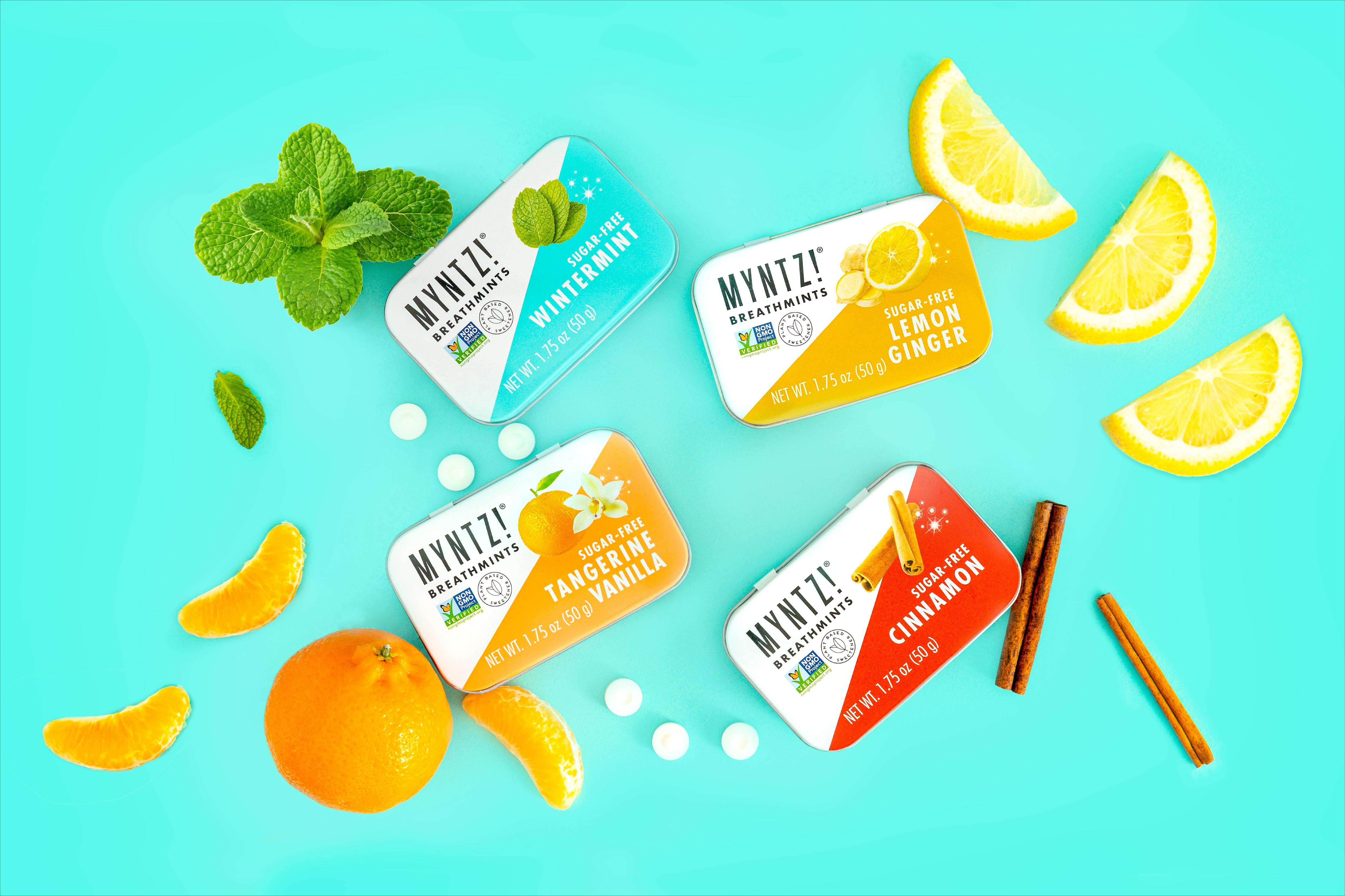 Four tins of MYNTZ! breathmints in different flavors with mint, lemon, tangerine and cinnamon with corresponding fruit and herbs.
