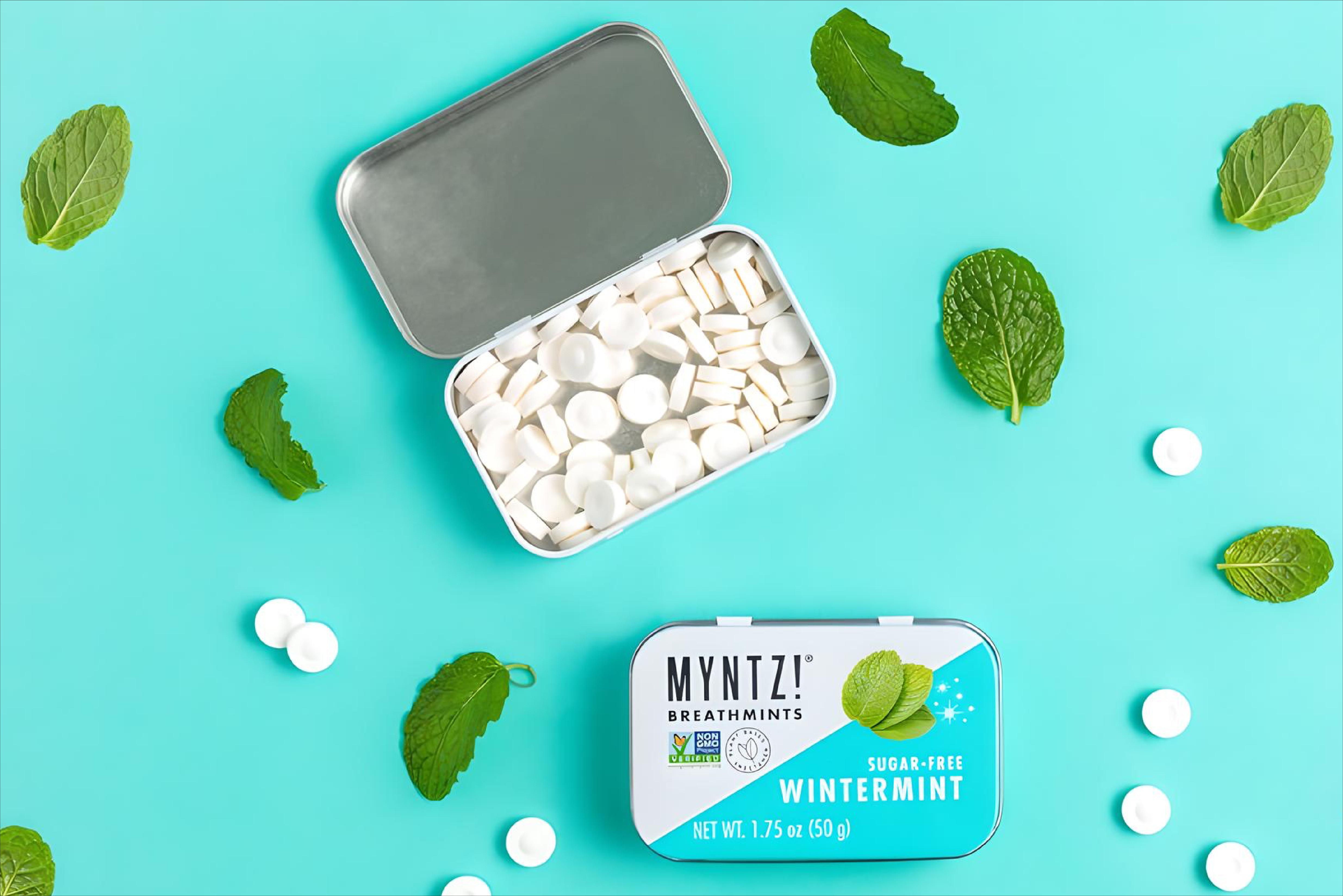 An open tin of MYNTZ! breathmints with an unopened tin and scattered mint leaves and mints on a light blue background.